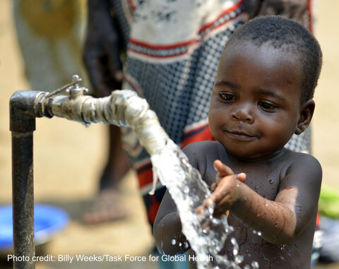 A young child in Malawi plays with water flowing from a tap, smiling as they hold their hands under the stream.